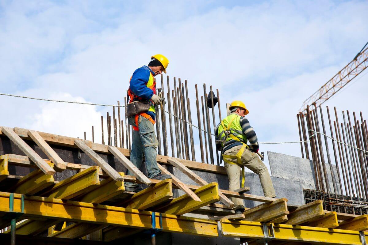 Dos operarios con casco, chaleco reflectante y arnés de seguridad caminan sobre un encofrado en una estructura de hormigón en construcción, con armaduras de acero y una grúa al fondo.